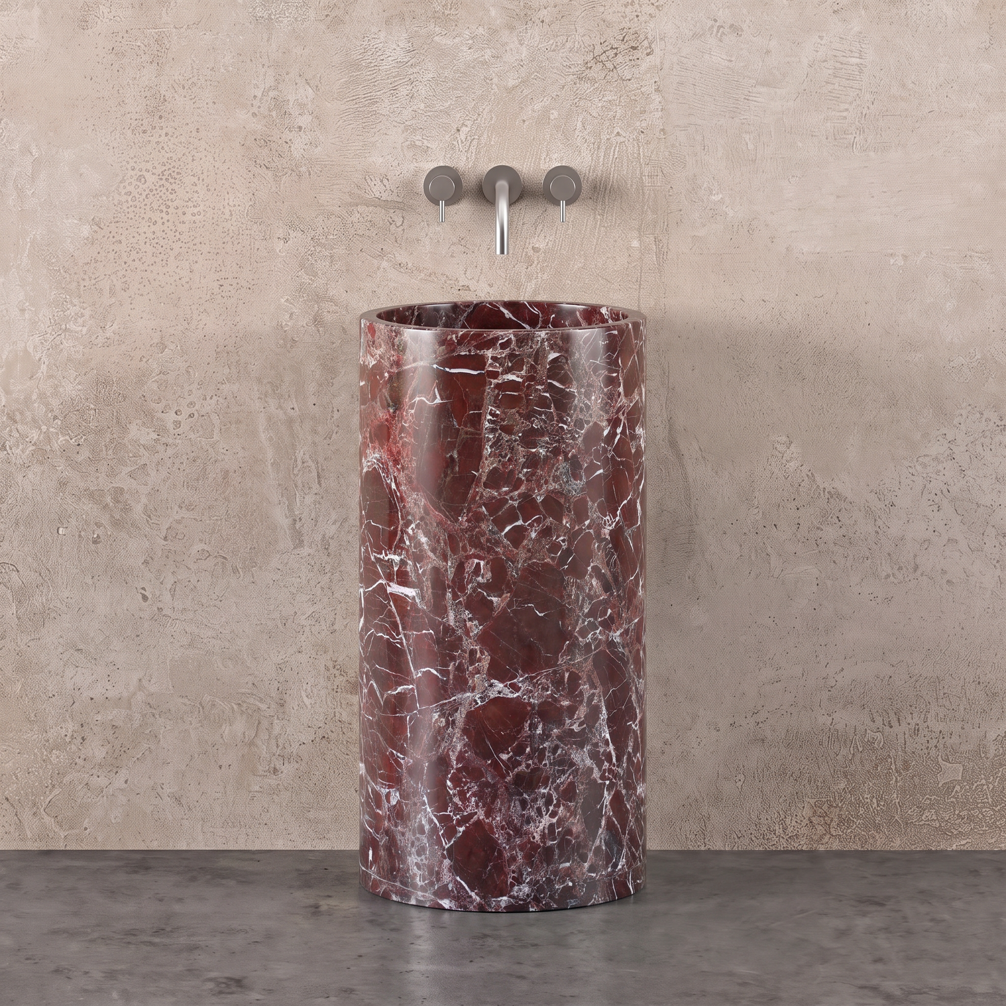 A tall cylindrical pedestal sink made of polished rosso levanto red marble with white veining, set against a textured beige plaster wall and dark gray concrete floor. A chrome three-hole faucet is mounted above the sink.